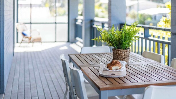 A wooden table with a plant and a croissant on it, set on a balcony with a view of the street. - Blue House on Pippi - Oceanstays - Pet Friendly (Yamba)
