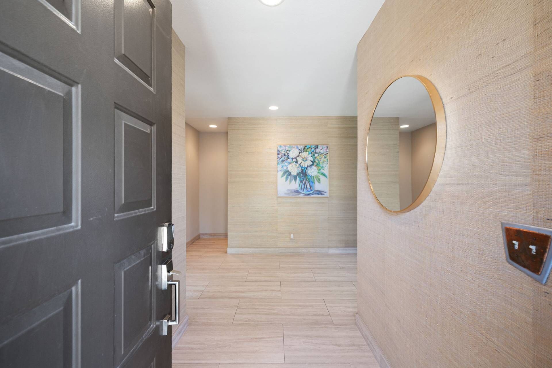 A hallway with a dark door, a light wooden wall, a painting, and a round mirror.