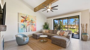 A spacious living room with a sectional sofa, a round coffee table, and a ceiling fan. There is a large window offering a view of palm trees and a building. - Palm Valley CC Charm | Outdoor Dining on the Patio, Desert Fun, Fitness, Spa, Tennis & Pickle Ball (Palm Desert)