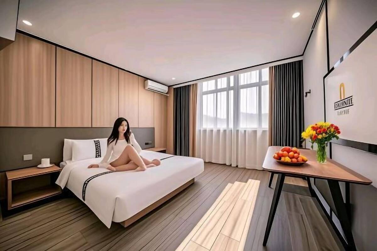 Una habitación de hotel moderna con una cama grande, una mesita de noche con un jarrón de flores y vistas a la ciudad a través de amplios ventanales.
