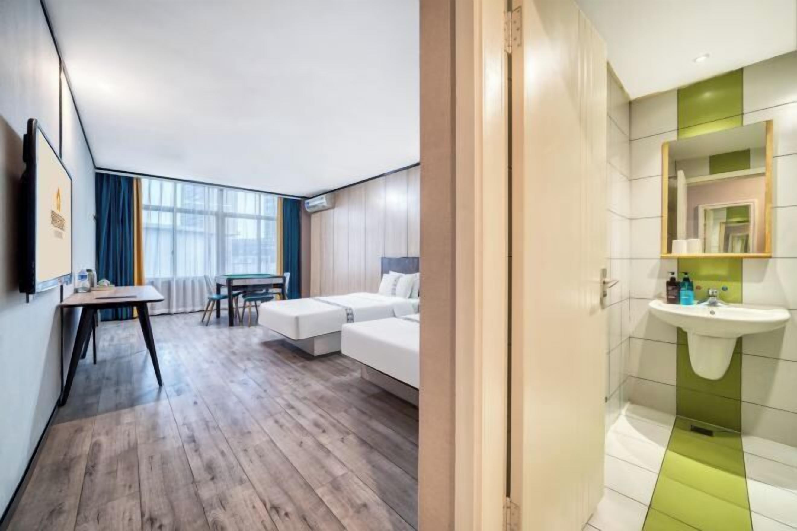 A hotel room with a wooden floor, a bed, a desk, and a bathroom with a green-tiled sink.