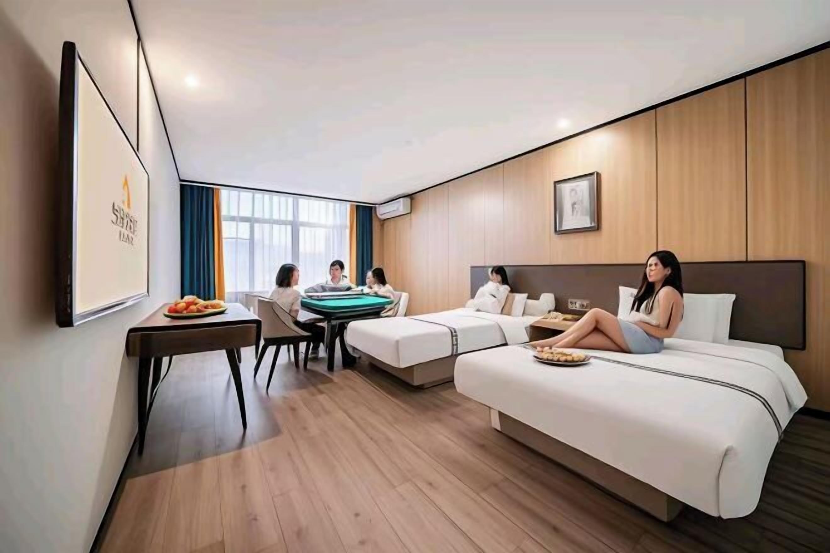 A modern hotel room with a large bed, a small sofa, a dining table with chairs, and a large window providing natural light.