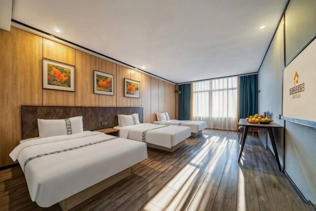 Habitación de hotel con tres camas, suelo de madera, un ventanal con cortinas y un frutero sobre la mesita de noche.
