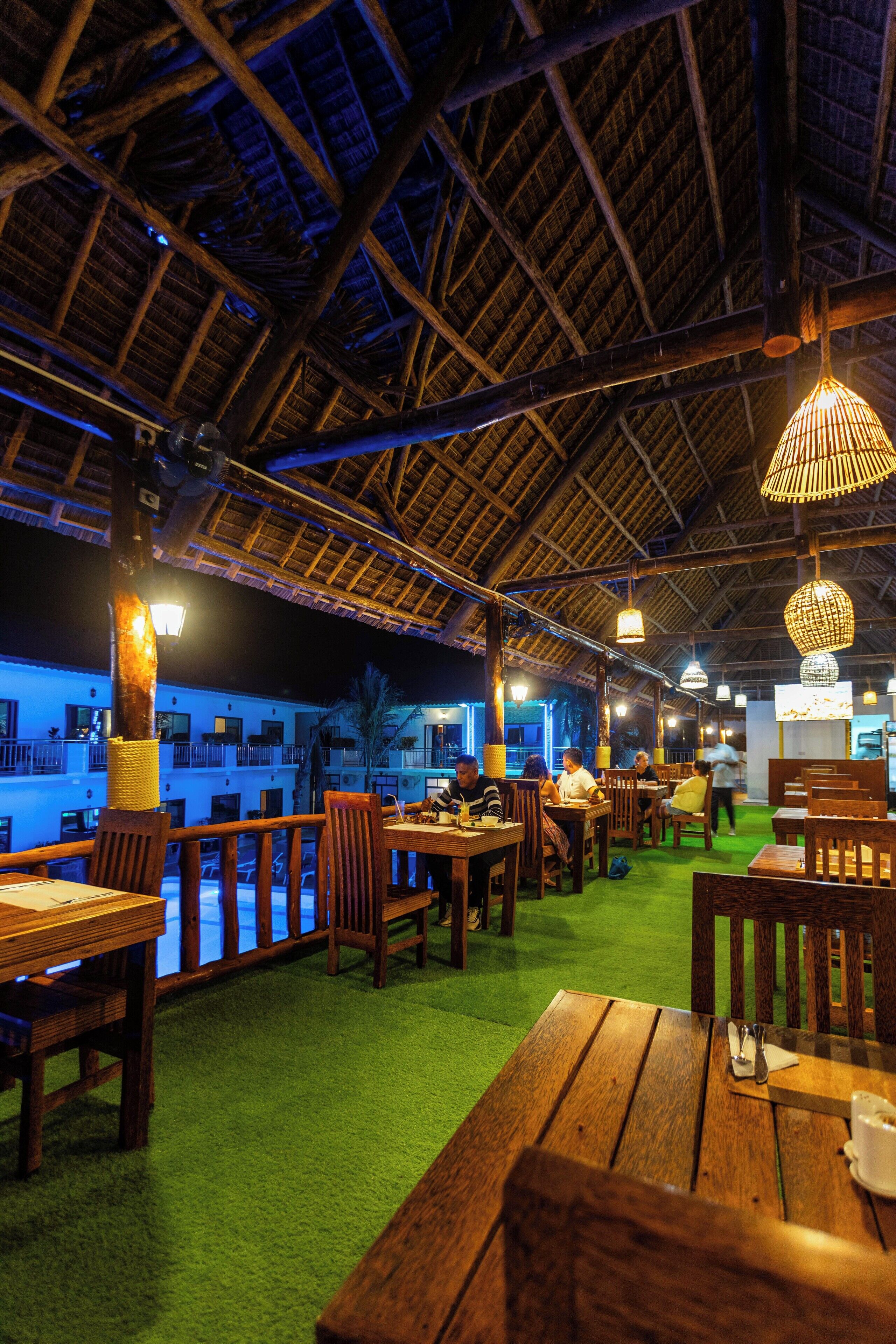 An outdoor dining area with wooden tables and chairs, a thatched roof, and hanging lights.