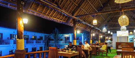 An outdoor dining area with wooden tables and chairs, a thatched roof, and hanging lights.
