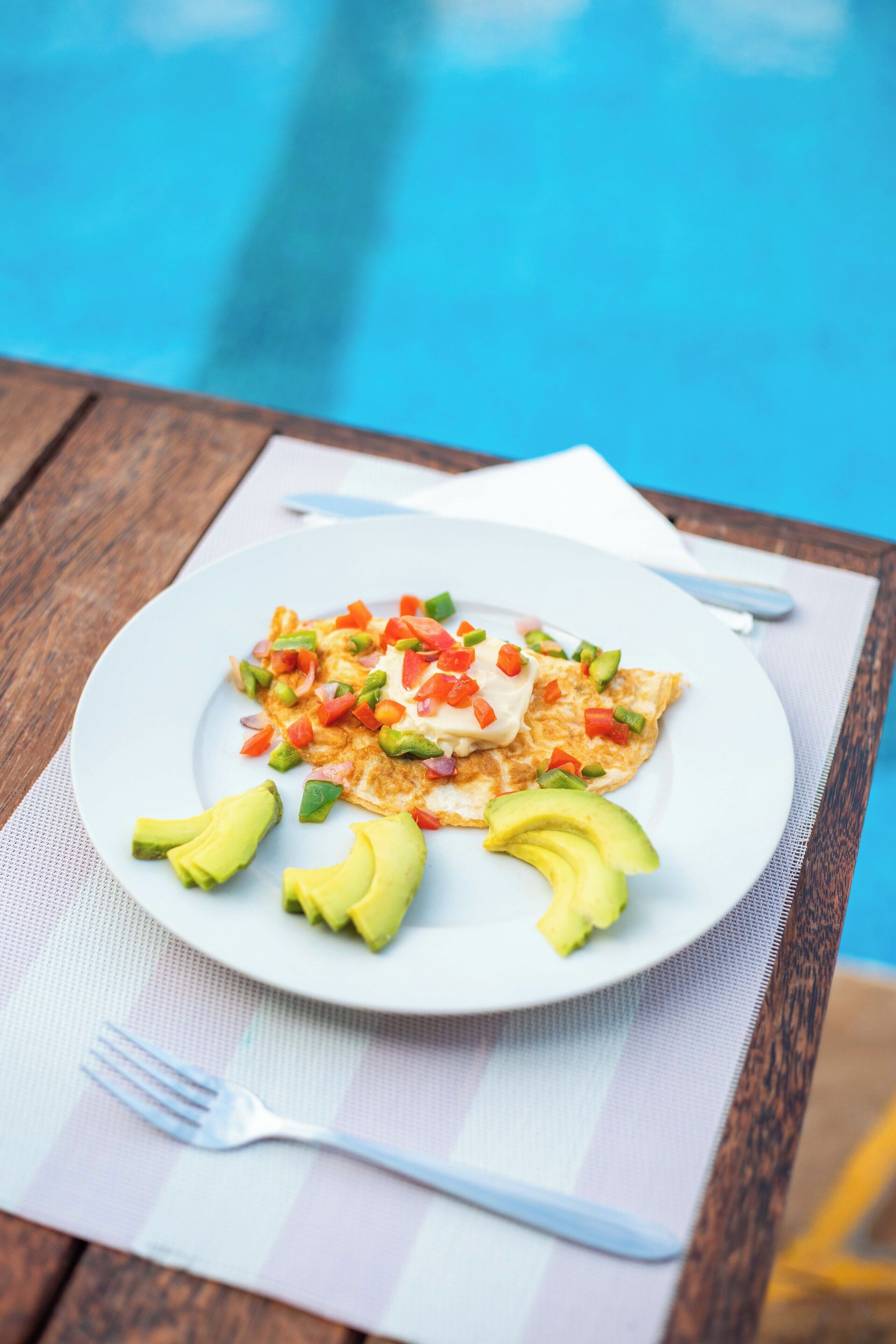 Un plato de comida con rebanadas de aguacate y verduras picadas, sobre un plato blanco, colocado en una mesa de madera con un tenedor y una servilleta a un lado.