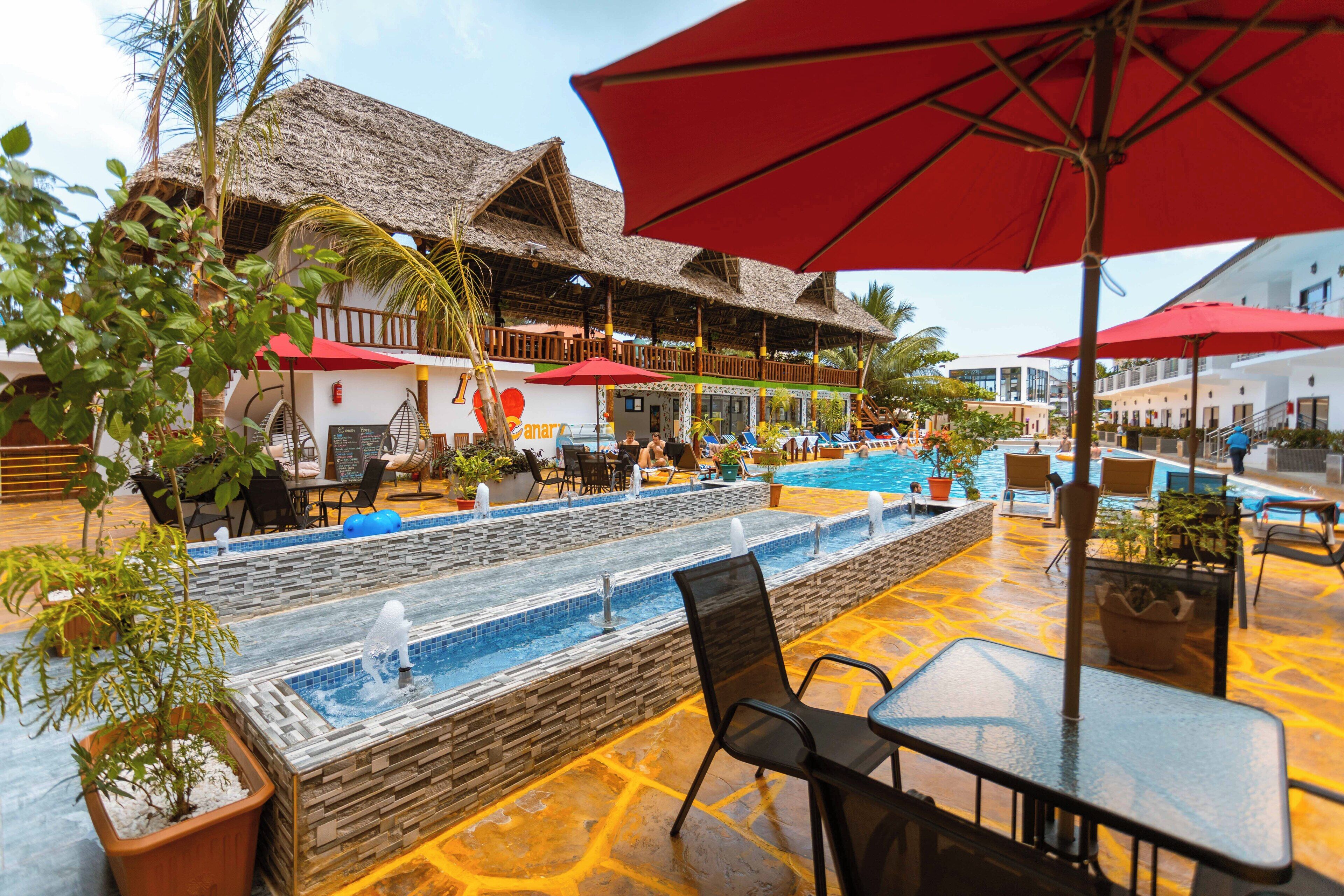 Un espace piscine agrémenté de chaises longues et de parasols, entouré de bâtiments aux toits de chaume, le tout dans un cadre tropical.