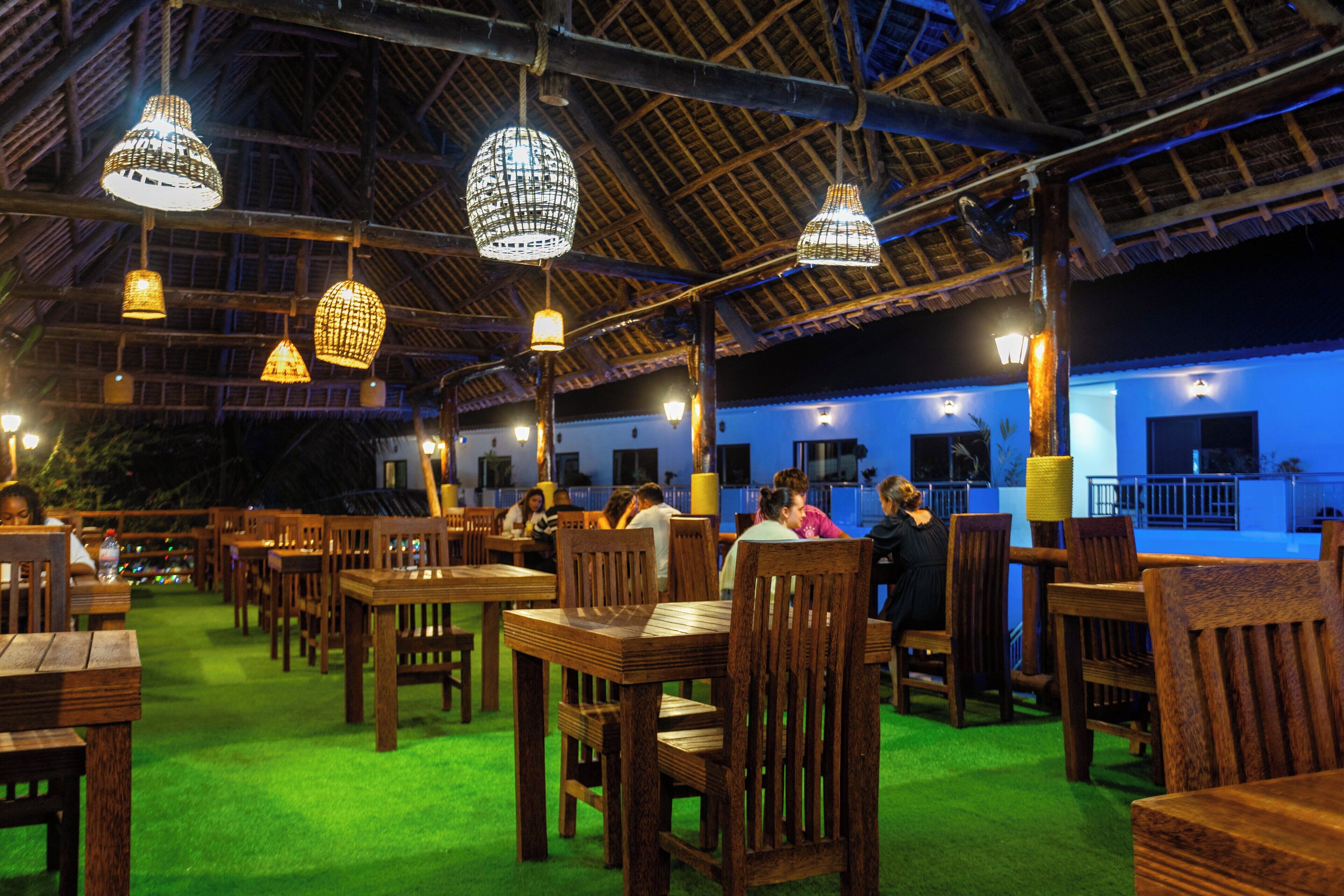 An indoor dining area with wooden tables and chairs, hanging light fixtures, and a green carpet.