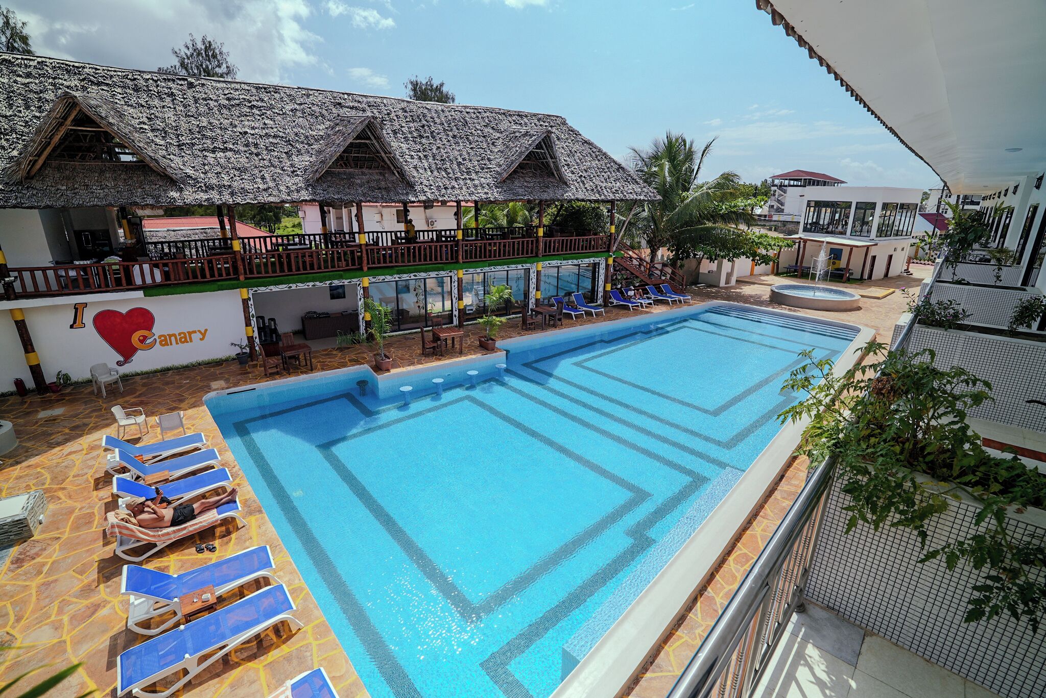 A swimming pool with lounge chairs, a covered area with a thatched roof, and a building with a 'I love Canary' sign.