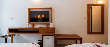 A hotel room with a bed, a desk with a television displaying 'NETFLIX', a small refrigerator, and a mirror on the wall.