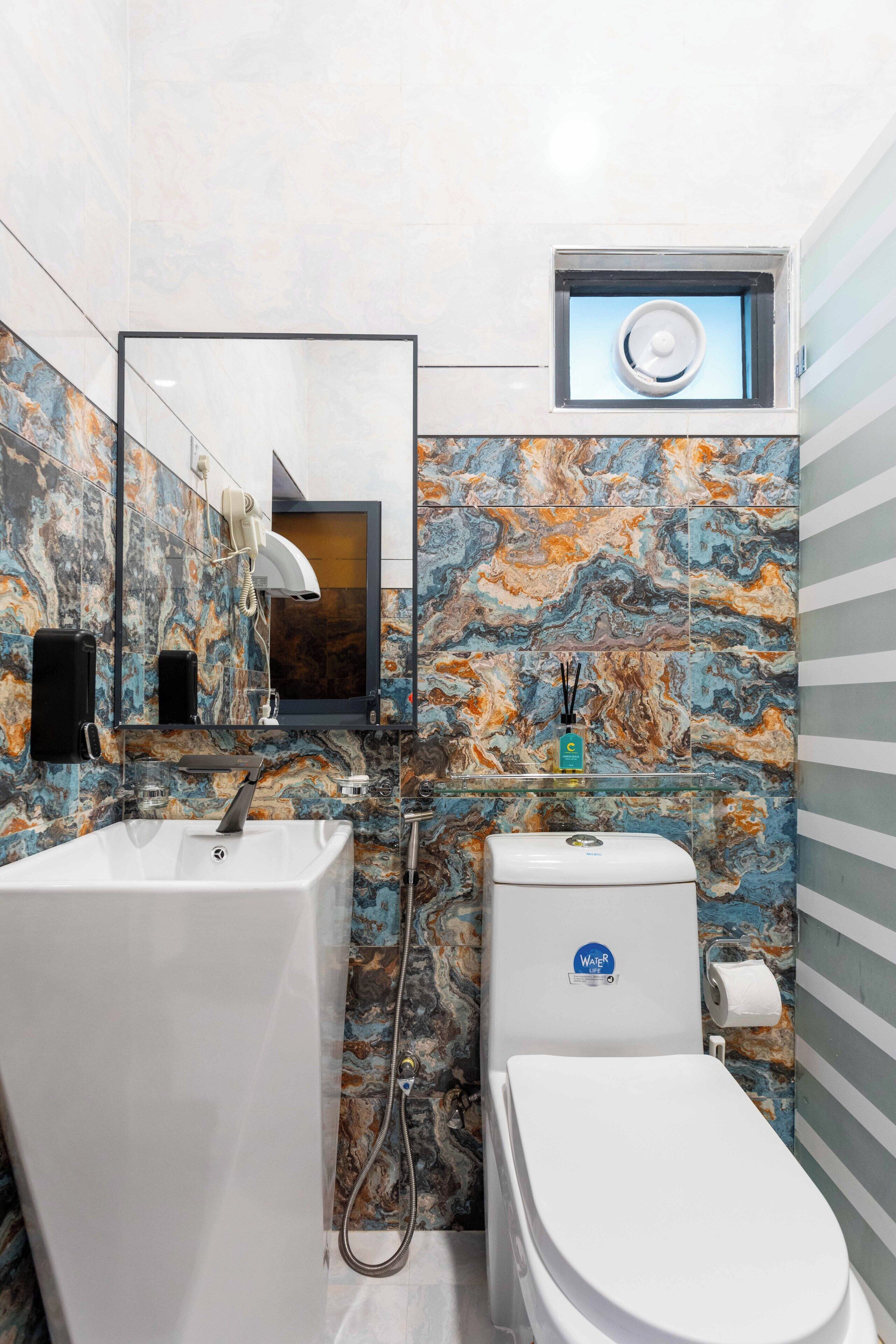 A bathroom with a marble backsplash, a white sink, a toilet, and a small window above the toilet.