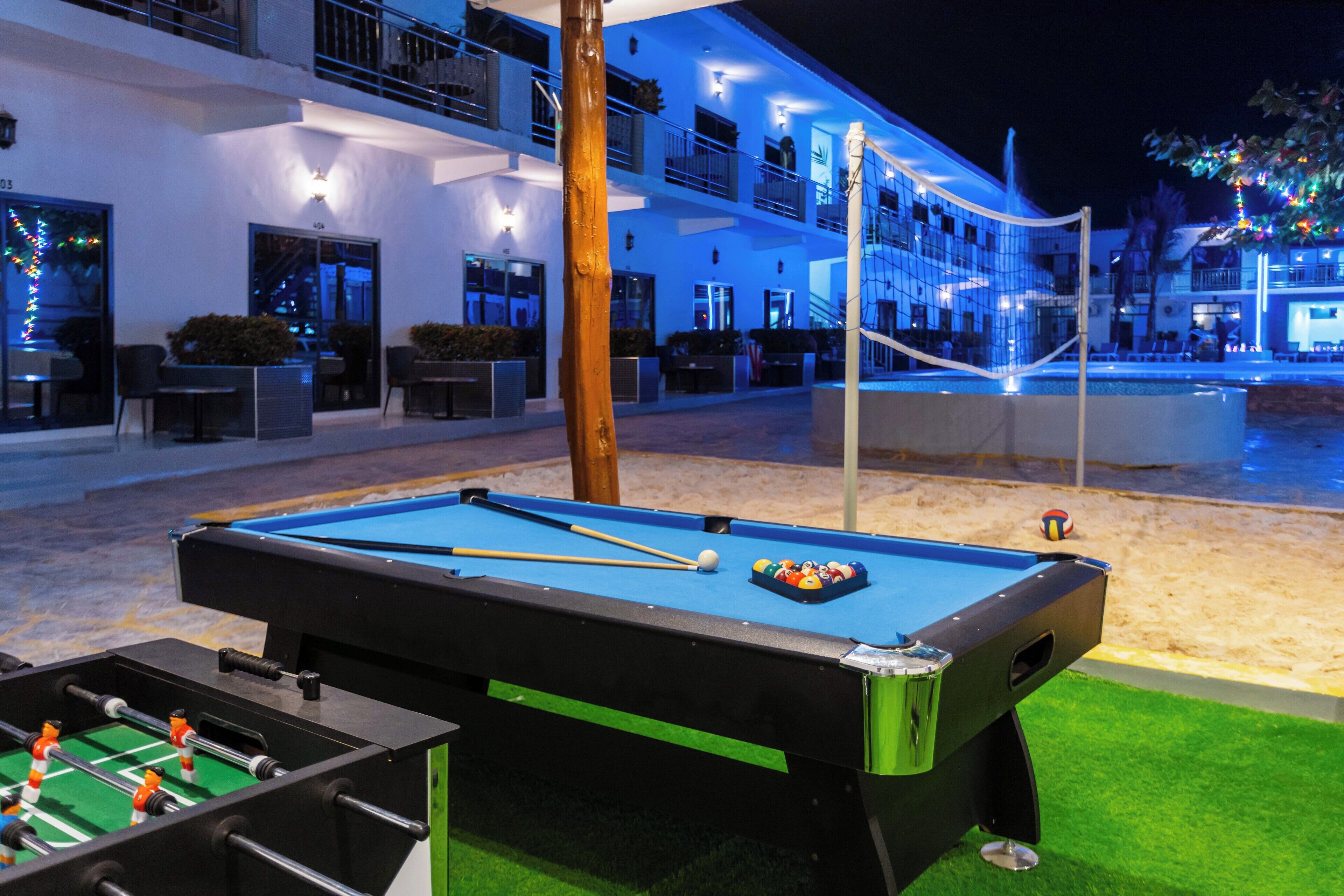 A pool table with a foosball table beside it, a tennis net, and a sandy area in the background.