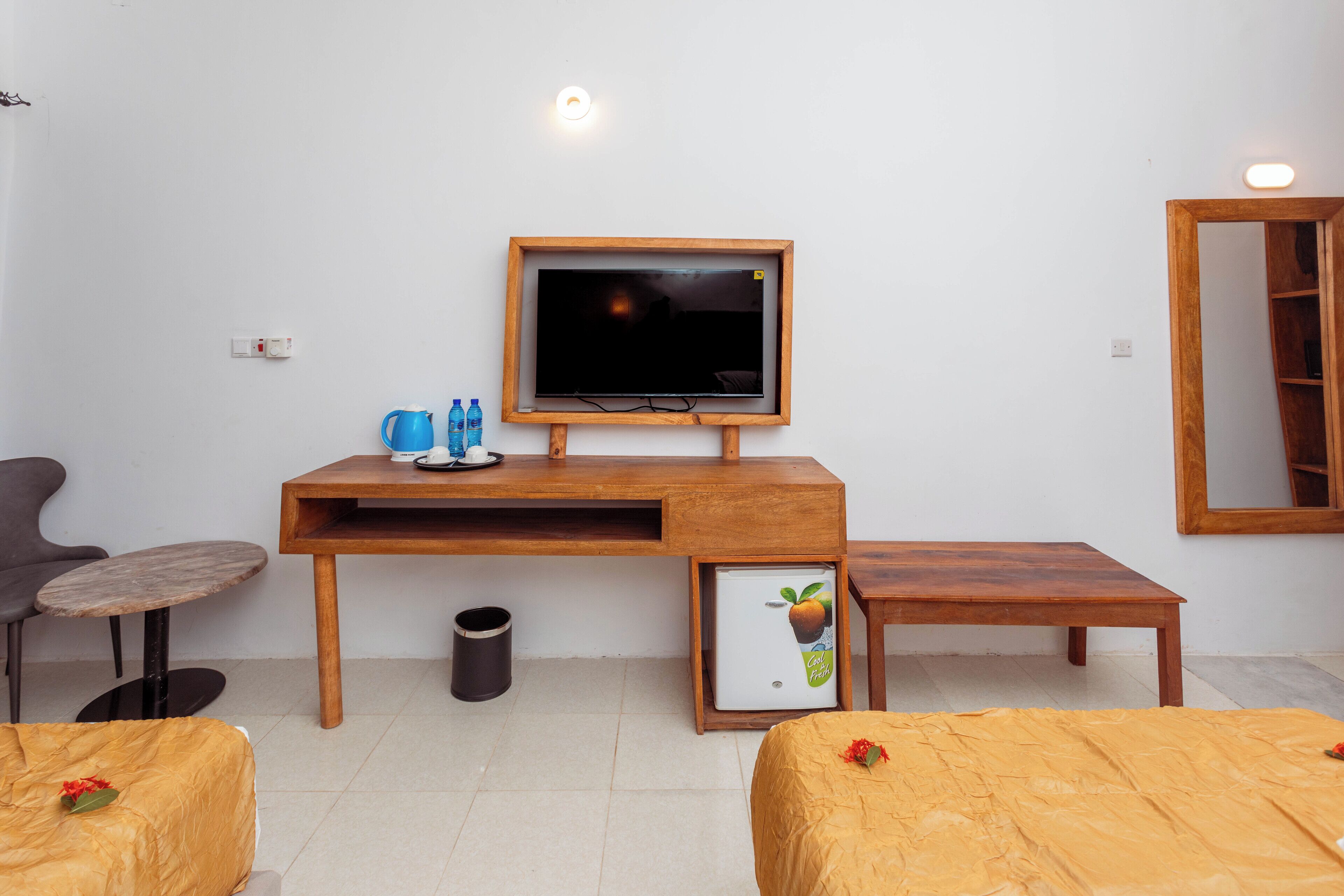 Ein Zimmer mit einem Holztisch, einem Fernseher, zwei Betten mit orangenen Decken, einem kleinen Tisch mit einer blauen Tasse und einem Mülleimer.