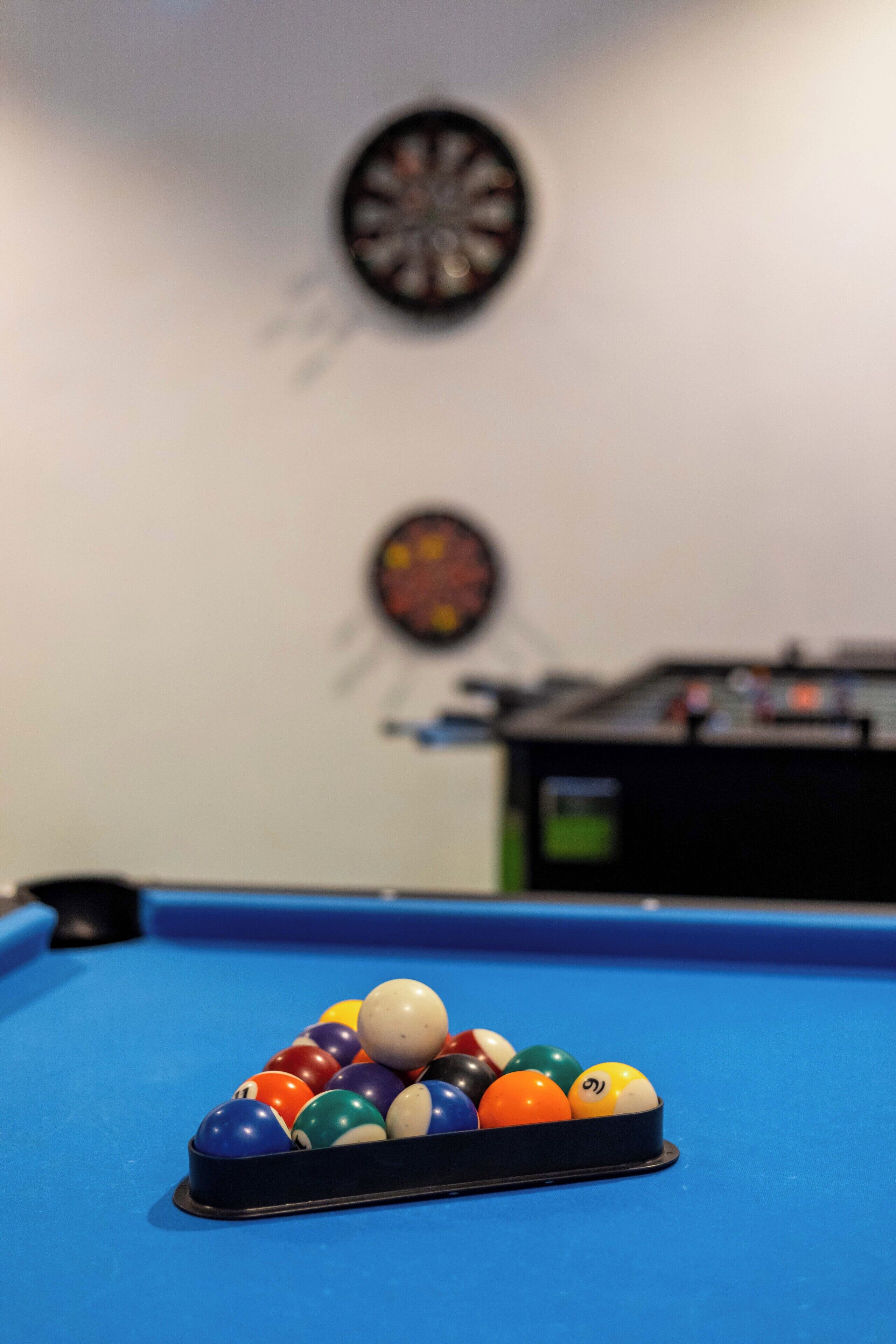 A pool table with a rack of billiard balls in a room with dartboards on the wall.