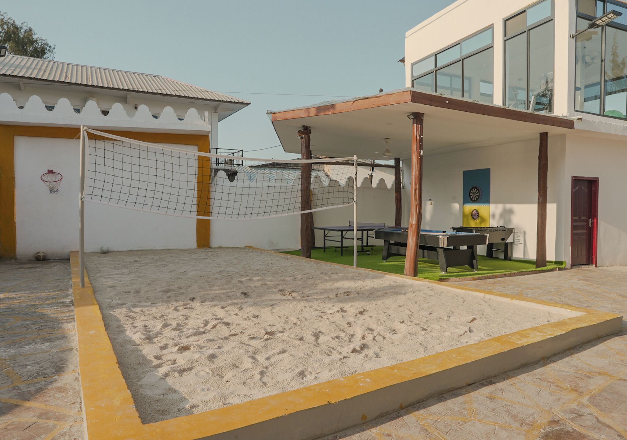 Una cancha de voleibol de arena con red, rodeada por un edificio con zona de estar cubierta y aro de baloncesto.