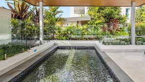 Et moderne poolområde udendørs med glasafskærmning, omgivet af frodigt grønt og en træpergola.