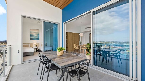 Et moderne udendørs spiseområde med et træbord og stole, udsigt til havet og en balkon med seng og skrivebord.