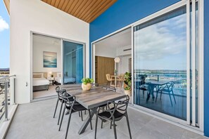 Een modern eetgedeelte buiten met een houten tafel en stoelen, uitzicht op zee, en een balkon met een bed en bureau.