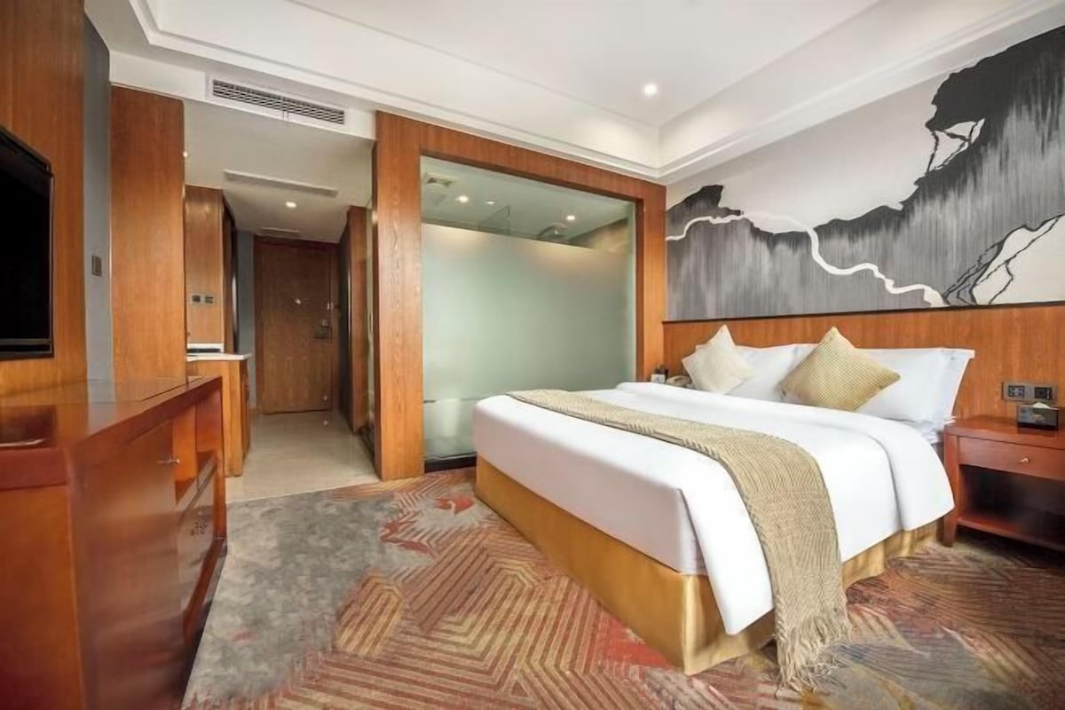 A hotel room with a large bed, a wooden dresser, a television, and a painting on the wall.