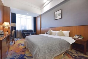 Une chambre d’hôtel avec un grand lit, un bureau avec une lampe, une chaise, une lampe de bureau, un téléphone et une fenêtre avec des rideaux.