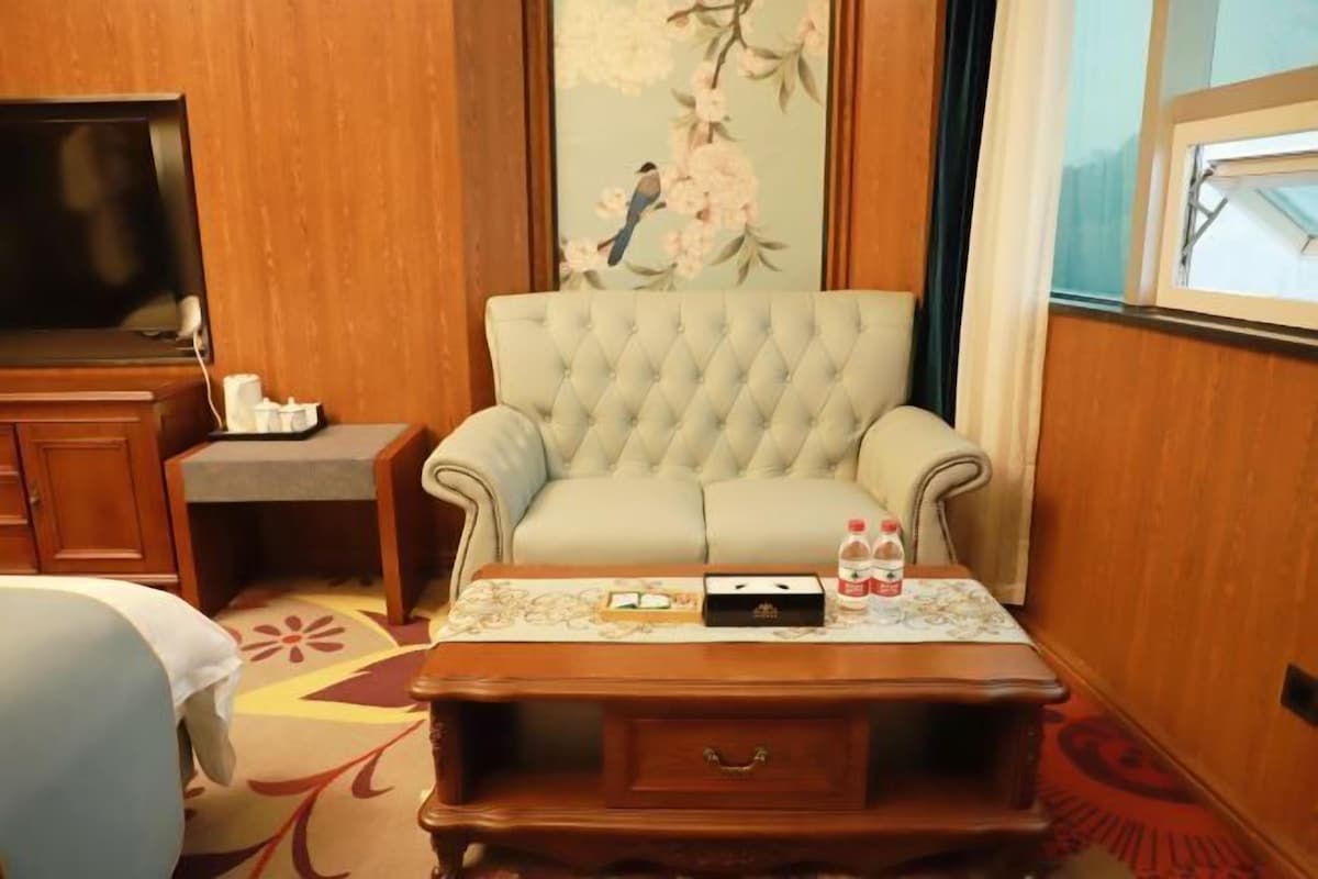 A room with a tufted armchair, a wooden side table with a remote control, a television, and a floral painting on the wall.