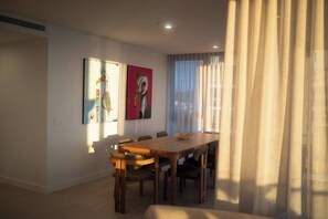 Ein Essbereich mit einem Holztisch und Stühlen, zwei an der Wand befestigten Kunstwerken und einem Fenster mit durchsichtigen Vorhängen.