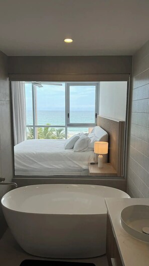 Ein Schlafzimmer mit großem Fenster mit Meerblick, einem Bett mit weißer Bettwäsche, einer Nachttischlampe und einer Badewanne.
