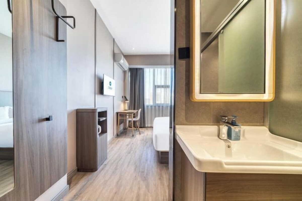 A modern hotel room with a wooden floor, a bed, a desk, and a bathroom with a sink and mirror.