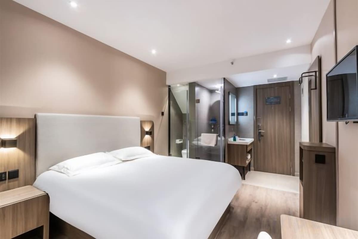 Ein modernes Hotelzimmer mit einem großen Bett, Nachttischen, einem Spiegel, einem Fernseher und einem Badezimmer mit Glasabtrennung.