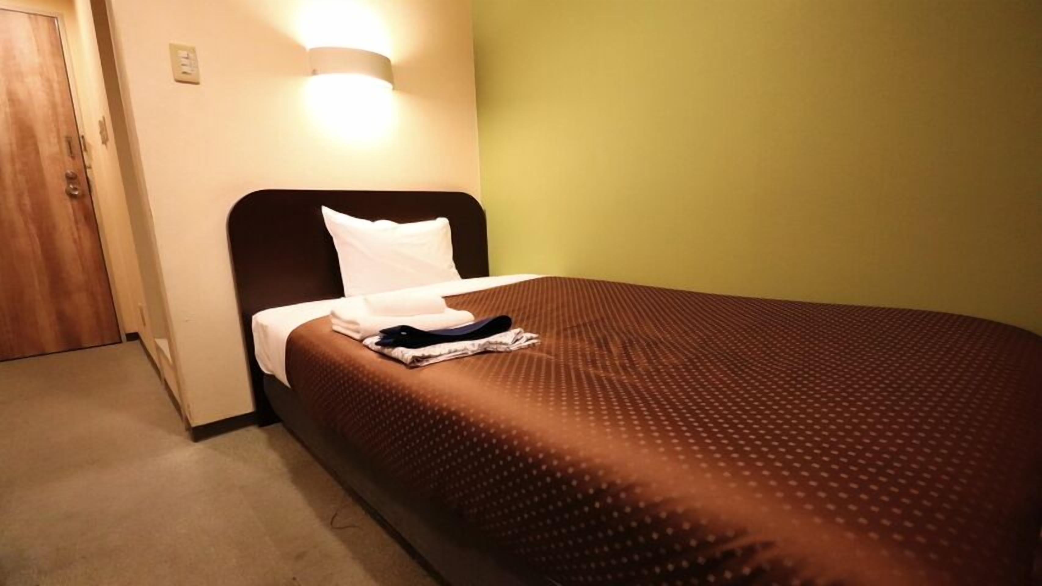 Een enkel bed met een bruine plaid en wit kussen in een kamer met een houten deur en een wandlamp.
