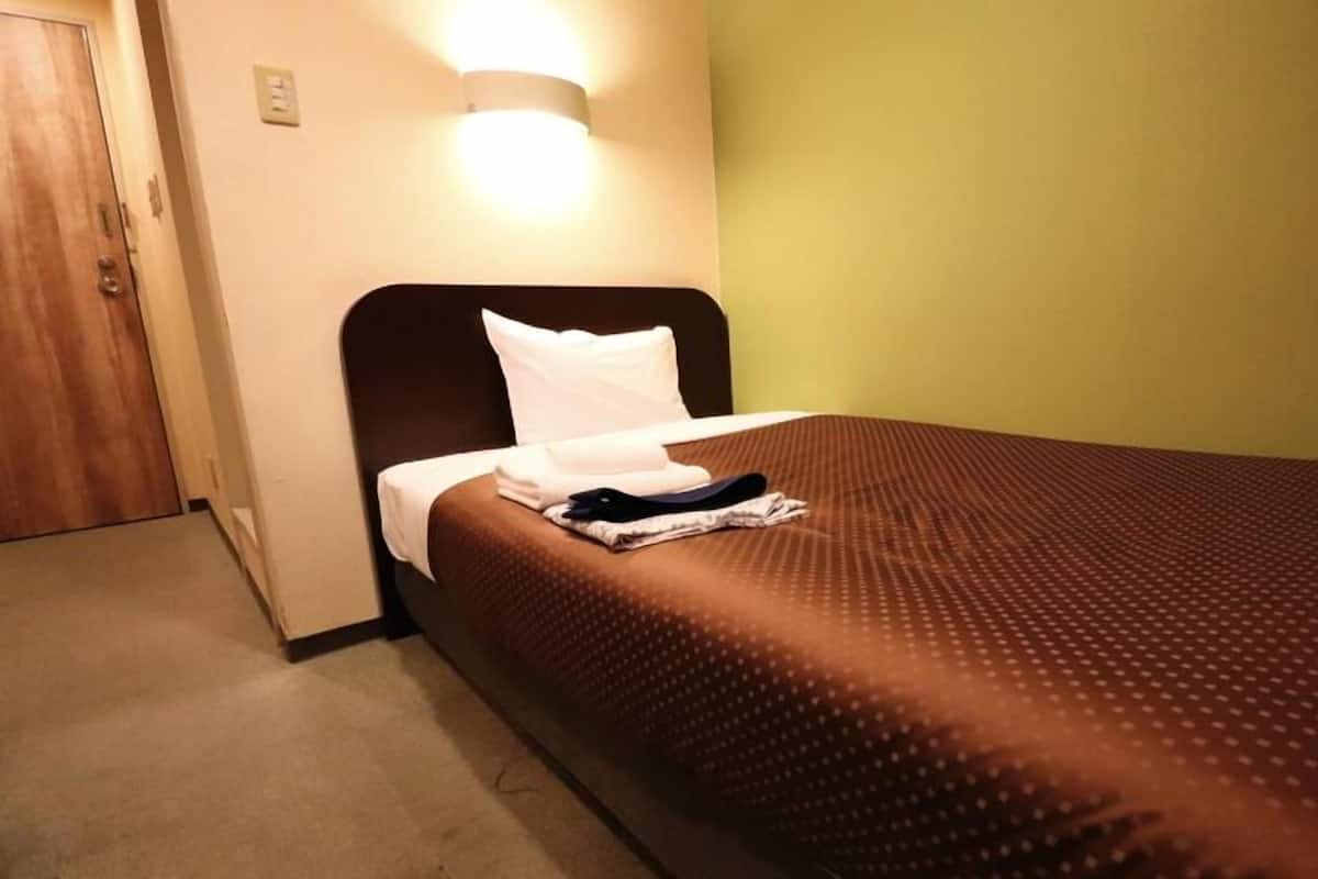 A single bed with a brown bedspread and white pillow in a room with a wooden door and a wall-mounted light fixture.