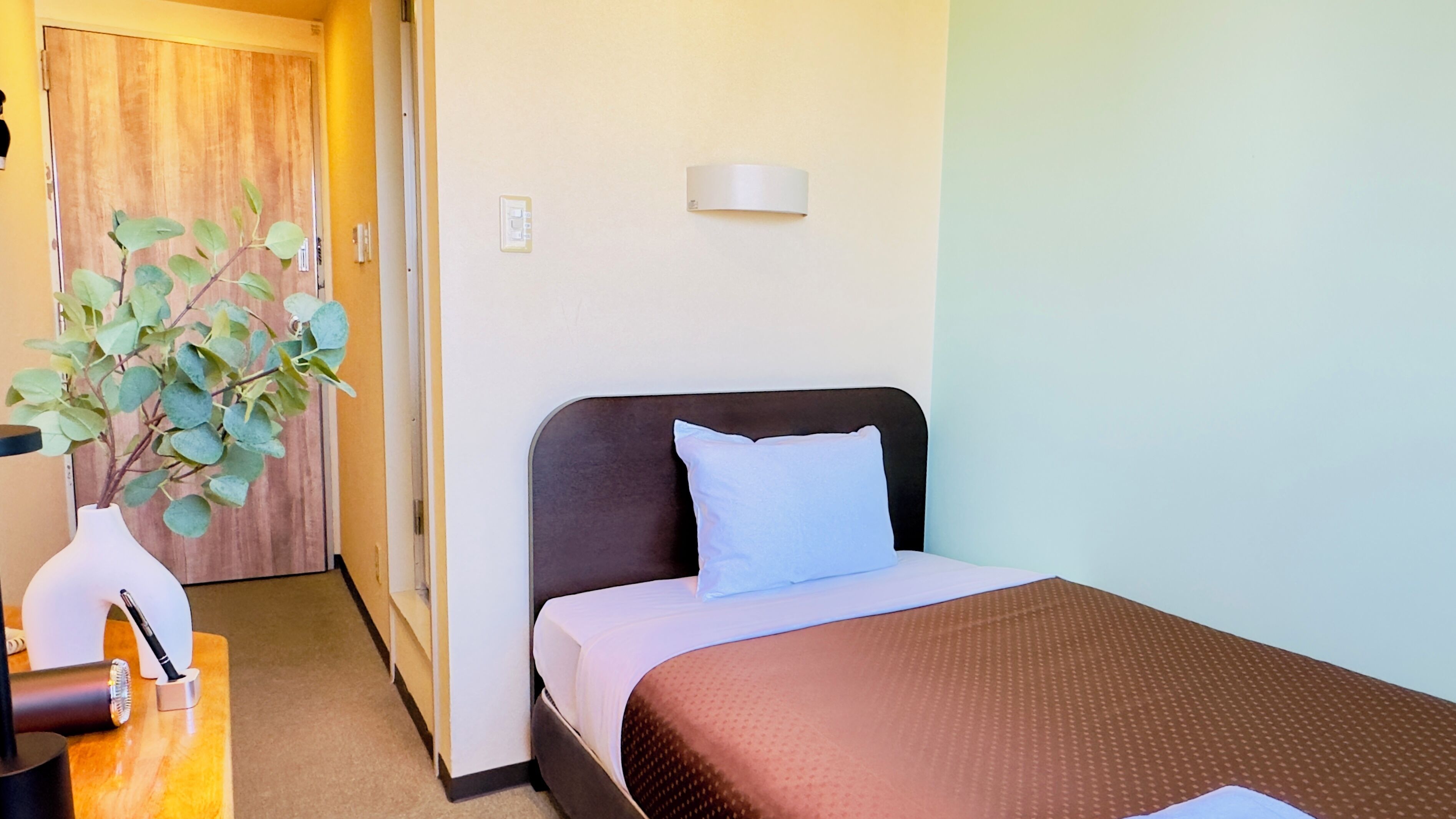 A hotel room with a bed, a bedside table with a pen holder, a wall-mounted light, and a door leading to another room.