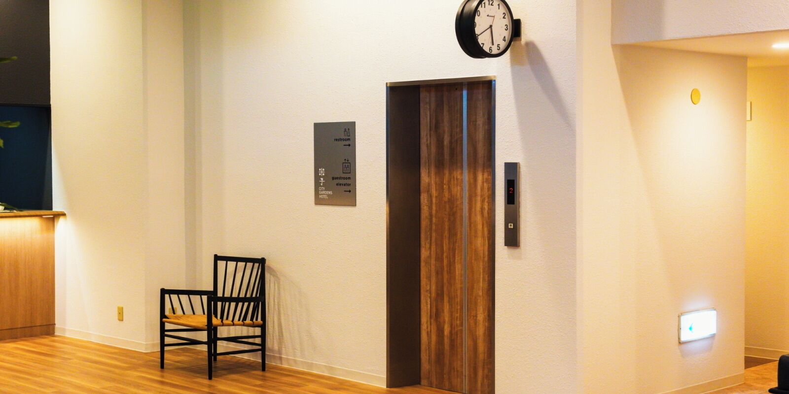A hotel lobby with a wooden floor, a black chair, a clock, and a door with a sign indicating the restroom and guest elevator.