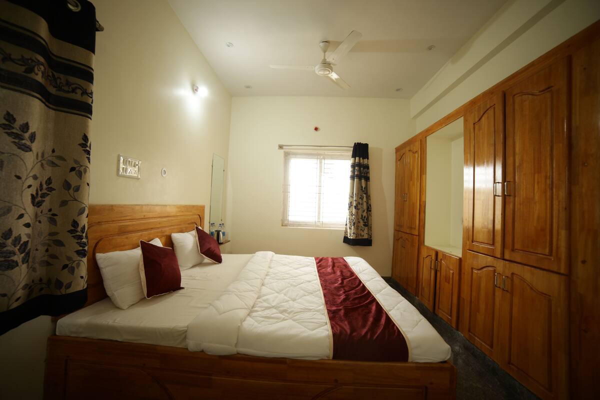 Ein Schlafzimmer mit Bett, Holzschrank, Deckenventilator, Fenster mit Vorhängen und einem an der Wand montierten Lichtschalter.