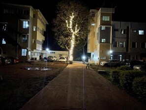 Un chemin menant à un immeuble à étages, entouré de voitures garées et d’un arbre décoré de guirlandes lumineuses.