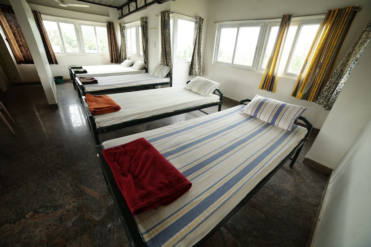 Ein Schlafsaalzimmer mit vier Einzelbetten, jeweils mit gestreifter Bettdecke und Kissen. Das Zimmer verfügt über große Fenster mit Vorhängen, einen Fliesenboden und ein schlichtes, funktionales Design.