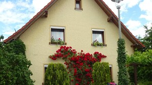 A house with a sloped roof, two windows, and a garden with red flowers and green shrubs. - Apartment 'Wassergartenparadies' with Shared Terrace, Shared Garden and Wi-Fi (Harsdorf)