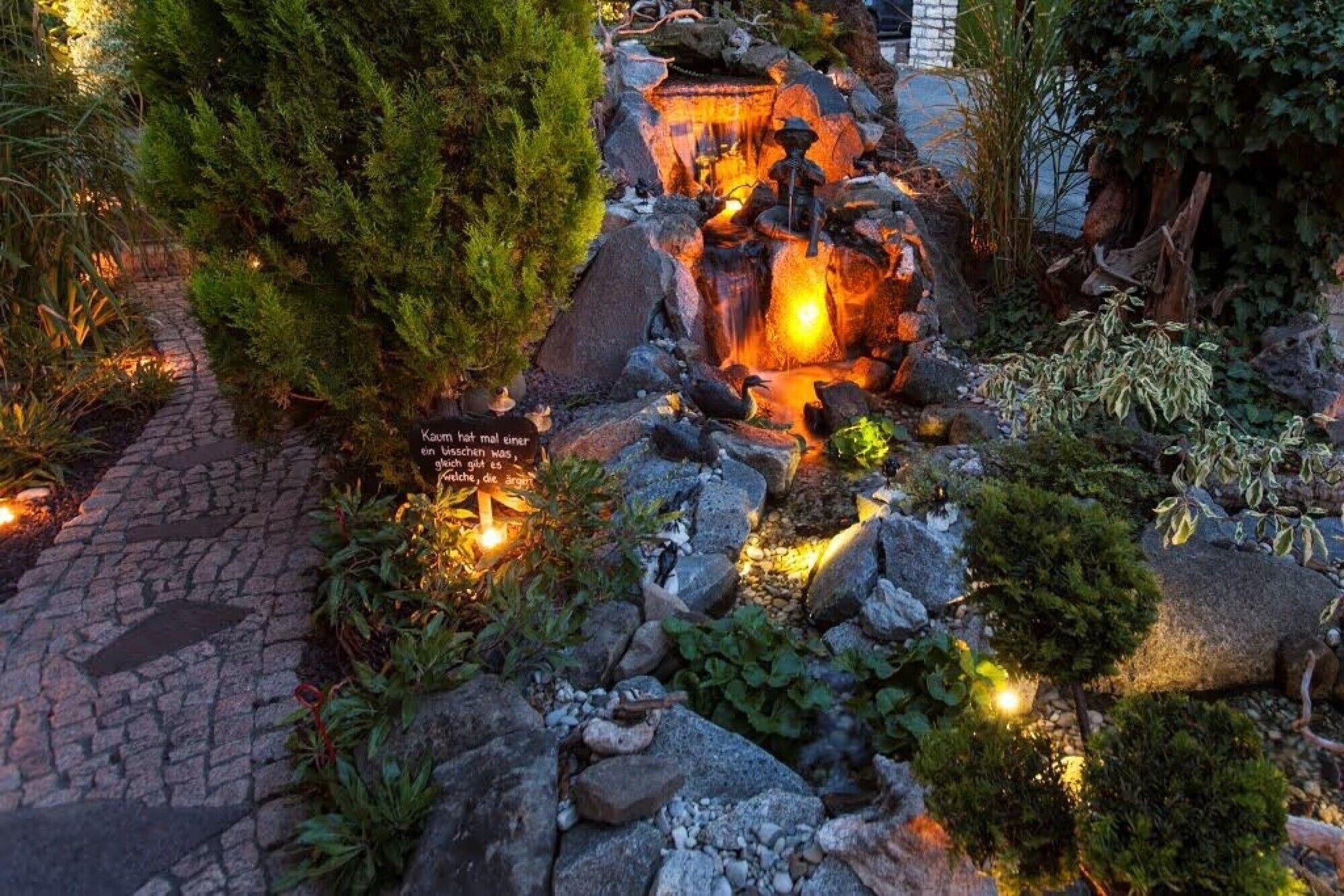 Un jardin avec un sentier en pierre, une petite cascade et un panneau avec du texte.