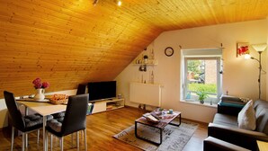 Una sala de estar acogedora con techo de madera, televisor, mesa de comedor con sillas, sofá y ventana con vista a follaje.