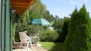 Une terrasse avec une chaise et une table, un parasol vert et une pelouse bien entretenue, agrémentée d’un grand arbre à feuilles persistantes.