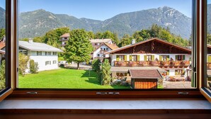 A view through a window of a building with a balcony, a green lawn, and mountains in the background. - Apartment 'Ferienwohnung Waxensteinblick' with Mountain View, Balcony and Wi-Fi (Grainau)