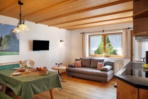 A cozy room with a wooden ceiling, a sofa, a dining table with a green tablecloth, a television, and a window with curtains.