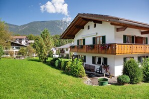 Un bâtiment à deux étages doté d’un balcon, entouré de verdure et avec des montagnes en arrière-plan.