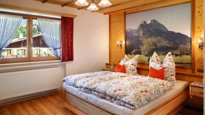 A bedroom with a wooden ceiling, a large bed with floral bedding, a window with curtains, and a mountain landscape painting on the wall. - Apartment 'Ferienwohnung Waxensteinblick' with Mountain View, Balcony and Wi-Fi (Grainau)