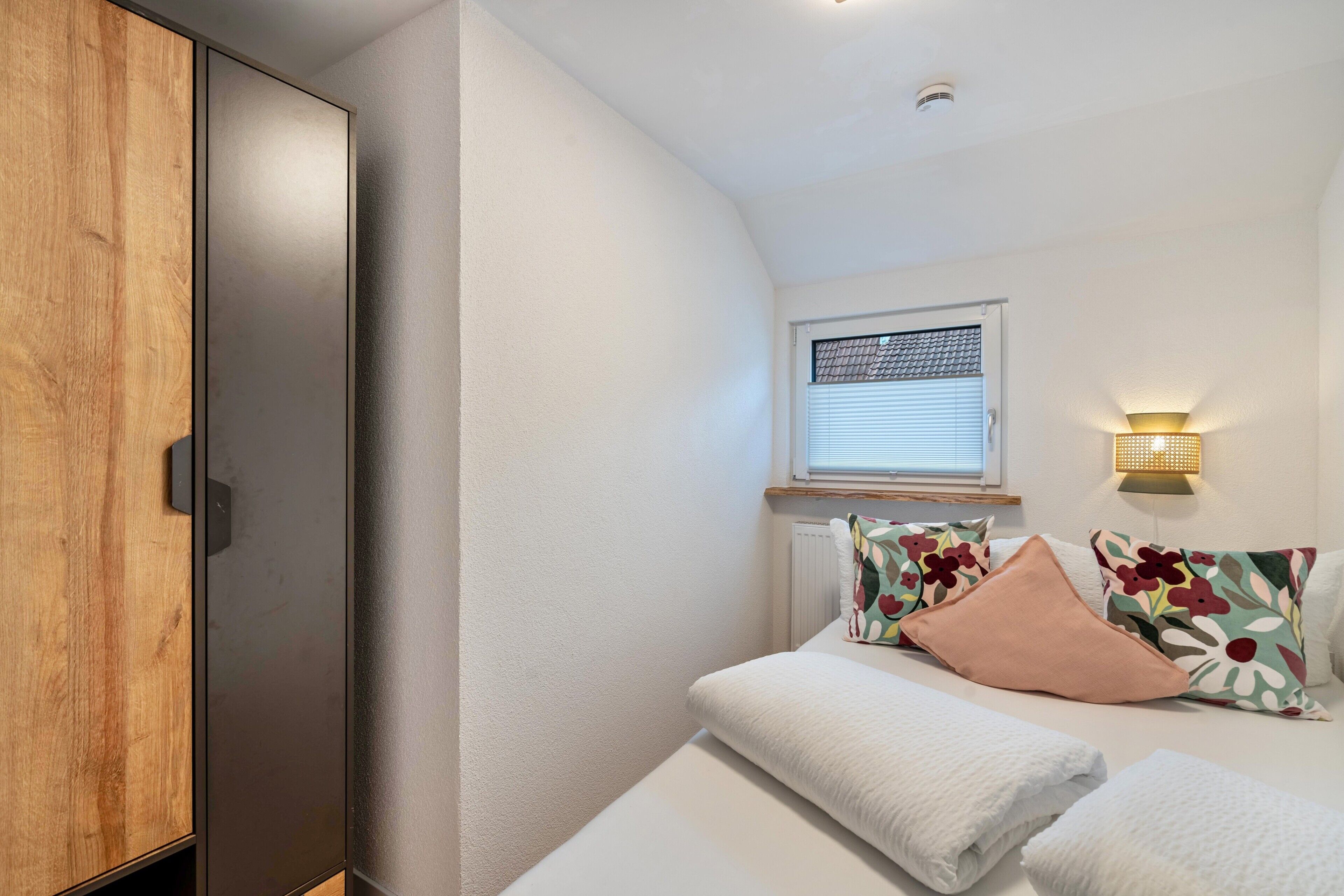 A bedroom with a wooden wardrobe, a bed with floral pillows, a bedside table, and a wall-mounted light.