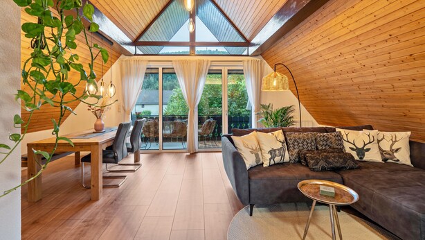 Una habitación acogedora con techo de madera, un sofá marrón oscuro con almohadones con motivos de ciervo, una mesa de comedor de madera y una planta colgante.