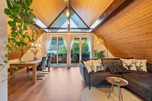 Una habitación acogedora con techo de madera, un sofá marrón oscuro con almohadones con motivos de ciervo, una mesa de comedor de madera y una planta colgante.