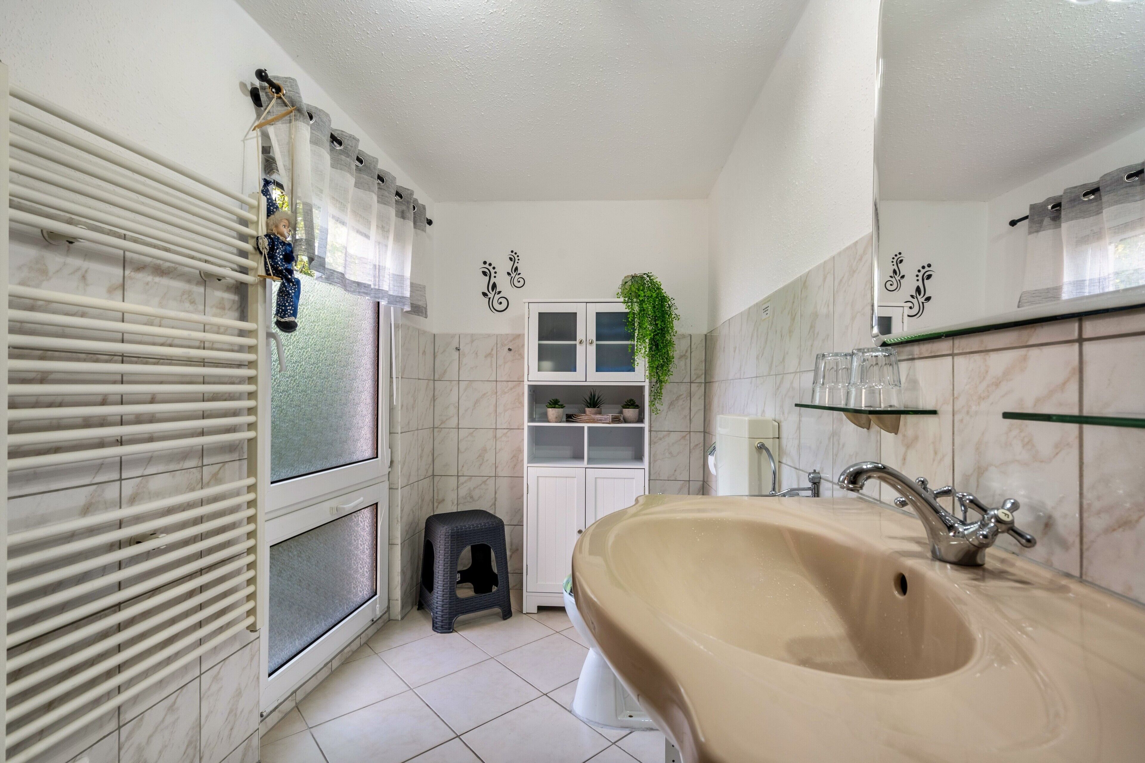 Ein Badezimmer mit einer großen Badewanne, einer Toilette, einem kleinen Hocker, einem Schrank mit Pflanzen und einem Fenster mit Jalousien.