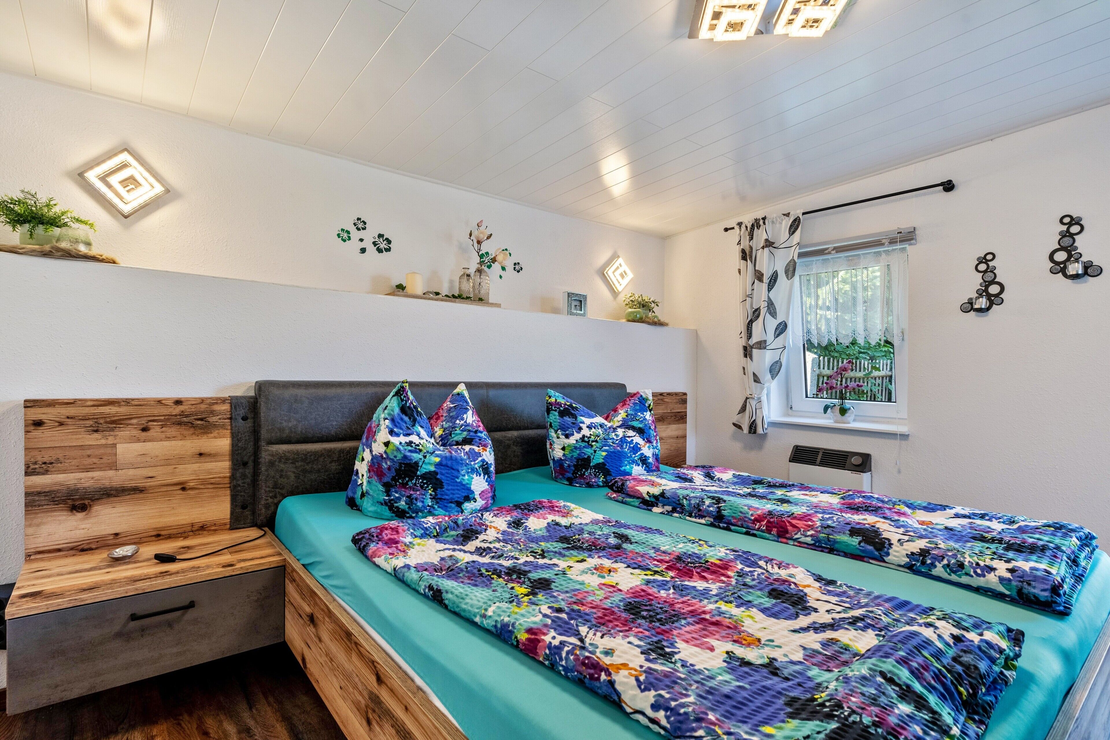 Une chambre moderne avec une tête de lit en bois, deux lits recouverts de couvertures colorées, une table de chevet et une fenêtre donnant sur un beau paysage.