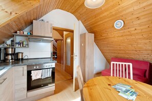 Uma cozinha com fogão, forno e máquina de café, uma mesa de jantar com revistas e um sofá vermelho.
